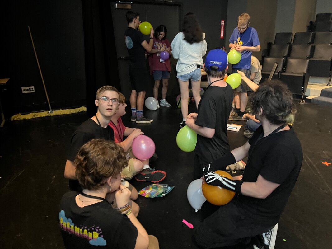 A bunch of kids working on a balloon tower with one staring directly at the camera.